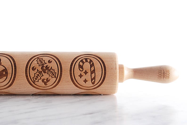 Wooden embossed rolling pin with festive holiday patterns placed on a light marble surface, with three freshly baked cookies in front showing detailed imprints such as holly and a snowflake. In the background, softly blurred Christmas lights and decorated trees create a warm golden bokeh effect. The scene highlights the natural wood texture, precise engraving, and cozy seasonal atmosphere, combining product detail with a festive, high-end lifestyle setting.