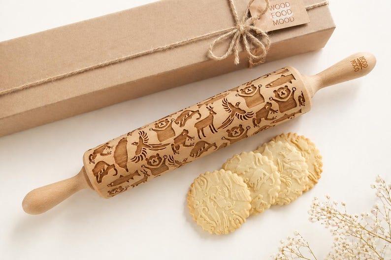 A beautifully styled product scene featuring a wooden embossed rolling pin with intricate animal patterns, placed diagonally on a soft, light background. Next to it lies a kraft gift box tied with natural twine, enhancing the rustic yet elegant aesthetic. In the foreground, several round cookies display detailed imprints from the rolling pin, showcasing real baking results. Delicate dried flowers add a refined, minimal touch, while soft lighting and gentle shadows create an inviting composition.