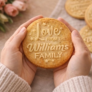 May include: A round, golden-brown cookie with the words "Love from the Williams Family" embossed on its surface. The cookie is held in two hands, with other cookies visible in the background. Pink flowers are in the background.