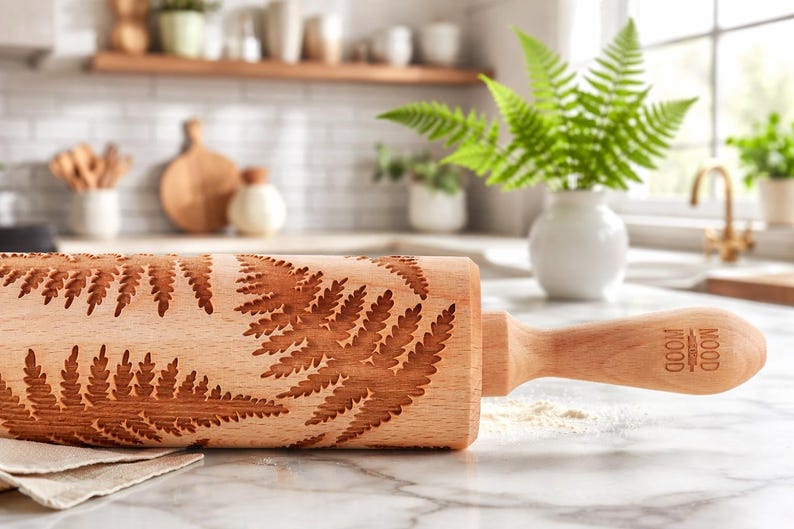 A wooden rolling pin with an engraved fern leaf pattern rests on a light marble countertop in a bright kitchen. In the background, a white vase with green fern leaves and light-colored kitchen furnishings create a cozy, natural atmosphere.