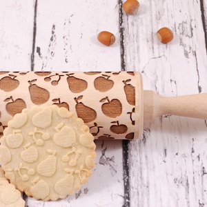 Wooden rolling pin with embossing design of apples, pears and cherries. Round cookiepresenting the results of embossing is standing in front of it.