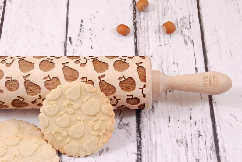 Wooden rolling pin with embossing design of apples, pears and cherries. Round cookiepresenting the results of embossing is standing in front of it.