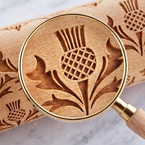 Close-up through a magnifying glass of a wooden rolling pin with a deep, approximately 3 mm engraved thistle pattern; the magnification highlights the sharp edges of the design and the wood texture.