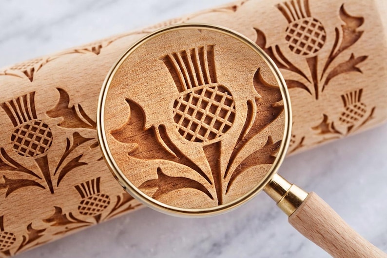 Close-up through a magnifying glass of a wooden rolling pin with a deep, approximately 3 mm engraved thistle pattern; the magnification highlights the sharp edges of the design and the wood texture.