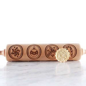 Wooden embossed rolling pin with festive holiday patterns placed on a light marble surface, with three freshly baked cookies in front showing detailed imprints such as holly and a snowflake. In the background, softly blurred Christmas lights and decorated trees create a warm golden bokeh effect. The scene highlights the natural wood texture, precise engraving, and cozy seasonal atmosphere, combining product detail with a festive, high-end lifestyle setting.