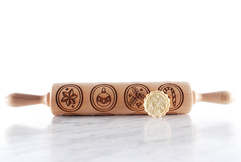 Wooden embossed rolling pin with festive holiday patterns placed on a light marble surface, with three freshly baked cookies in front showing detailed imprints such as holly and a snowflake. In the background, softly blurred Christmas lights and decorated trees create a warm golden bokeh effect. The scene highlights the natural wood texture, precise engraving, and cozy seasonal atmosphere, combining product detail with a festive, high-end lifestyle setting.