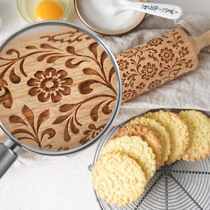 Close-up of an engraved wooden rolling pin with a floral pattern, styled on a light baking surface with cookies, flour, and ingredients. A realistic magnifying glass reveals a zoomed-in view of the exact same design, highlighting fine wood grain and deep 3 mm engraving detail. Elegant, minimal composition emphasizes craftsmanship, making it perfect for custom rolling pins, embossed cookies, baking tools, personalized gifts, and decorative baking ideas for Etsy listings.