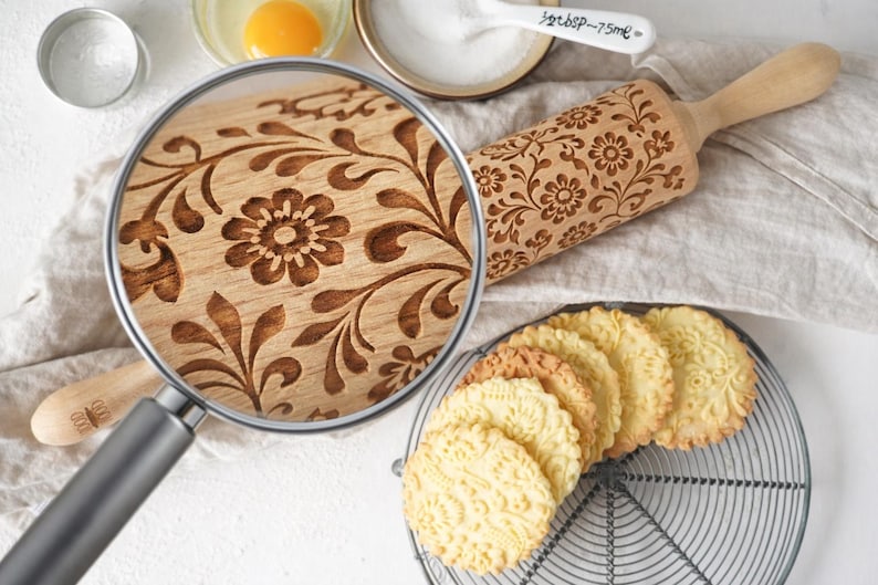 Close-up of an engraved wooden rolling pin with a floral pattern, styled on a light baking surface with cookies, flour, and ingredients. A realistic magnifying glass reveals a zoomed-in view of the exact same design, highlighting fine wood grain and deep 3 mm engraving detail. Elegant, minimal composition emphasizes craftsmanship, making it perfect for custom rolling pins, embossed cookies, baking tools, personalized gifts, and decorative baking ideas for Etsy listings.