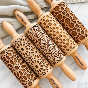 May include: Four wooden rolling pins with engraved geometric patterns and the brand name MoodForWood stamped on the handles. Each cylinder features a distinct, intricate design carved into the light-coloured wood surface for embossing dough.