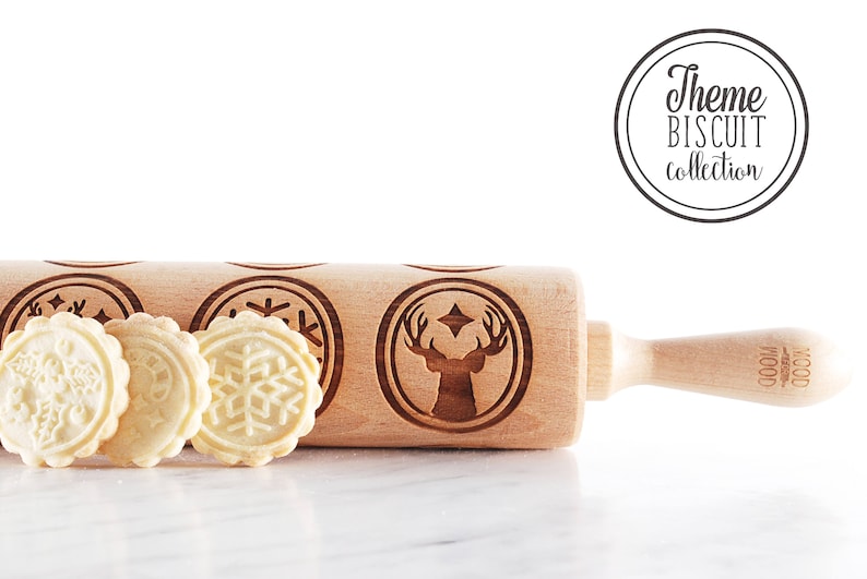 Wooden embossed rolling pin with festive holiday patterns placed on a light marble surface, with three freshly baked cookies in front showing detailed imprints such as holly and a snowflake. In the background, softly blurred Christmas lights and decorated trees create a warm golden bokeh effect. The scene highlights the natural wood texture, precise engraving, and cozy seasonal atmosphere, combining product detail with a festive, high-end lifestyle setting.