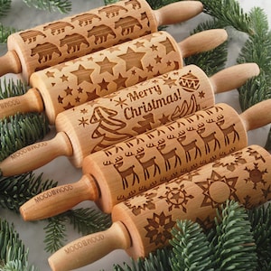 Five small wooden rolling pins with laser engraed designs on them: snowflakes, reindeer, merry christmas text, stars and polar bears. Among christmas tree branches.