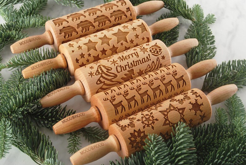 Five small wooden rolling pins with laser engraed designs on them: snowflakes, reindeer, merry christmas text, stars and polar bears. Among christmas tree branches.