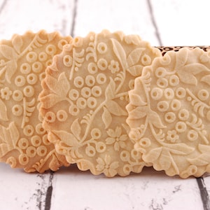 Puede incluir: Tres galletas redondas de color marr&oacute;n claro con un dise&ntilde;o floral en relieve en la superficie. Las galletas est&aacute;n sobre una superficie de madera blanca.