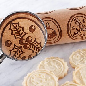 Wooden embossed rolling pin with intricate holiday patterns placed on a light marble surface, accompanied by freshly baked cookies. A realistic magnifying glass overlays the roller, revealing a perfectly matching enlarged view of the holly engraving, highlighting the natural wood grain and approximately 3 mm deep carving. The composition emphasizes precision, texture, and craftsmanship in a clean, high-end product photography style.