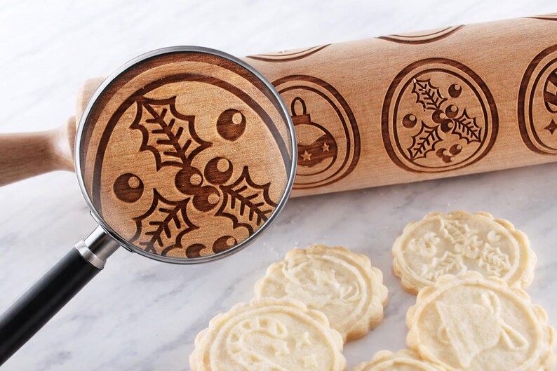 Wooden embossed rolling pin with intricate holiday patterns placed on a light marble surface, accompanied by freshly baked cookies. A realistic magnifying glass overlays the roller, revealing a perfectly matching enlarged view of the holly engraving, highlighting the natural wood grain and approximately 3 mm deep carving. The composition emphasizes precision, texture, and craftsmanship in a clean, high-end product photography style.