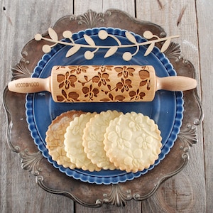 May include: A wooden rolling pin with an orchid design is shown on a blue plate with four cookies that have the same design. The rolling pin is labeled "WOODGOOD".