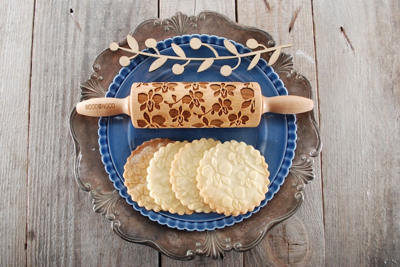 May include: A wooden rolling pin with an orchid design is shown on a blue plate with four cookies that have the same design. The rolling pin is labeled "WOODGOOD".