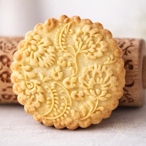 Close-up macro photograph of a single embossed butter cookie in warm golden tones, filling most of the frame and sharply focused to highlight intricate floral patterns and crisp edges. The cookie is positioned slightly off-center with strong contrast against a clean, softly blurred neutral background. A wooden engraved rolling pin sits behind it, out of focus but recognizable. Bright, soft natural lighting enhances texture and depth, creating a warm, handcrafted aesthetic