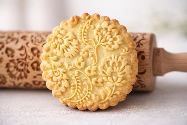 Close-up macro photograph of a single embossed butter cookie in warm golden tones, filling most of the frame and sharply focused to highlight intricate floral patterns and crisp edges. The cookie is positioned slightly off-center with strong contrast against a clean, softly blurred neutral background. A wooden engraved rolling pin sits behind it, out of focus but recognizable. Bright, soft natural lighting enhances texture and depth, creating a warm, handcrafted aesthetic