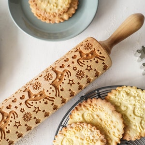 Wooden rolling pin for embossing cookies with reindeer design. Several well baked cookies are next to it onmetal trivet and plate. Steel cookie cutter in shape of snowflake is next to it.