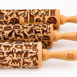 Puede incluir: Tres rodillos de madera de color claro con patrones grabados. El rodillo superior presenta animales de safari, el central muestra varias razas de perros con el texto "Woof" y "Hau", y el inferior muestra gatos. Cada mango tiene la marca "MOOD".
