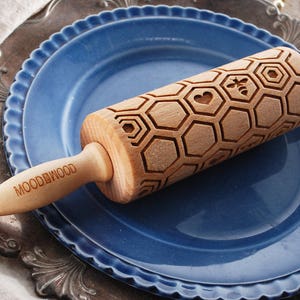 Wooden small rolling pin laser engraved with design of honeycomb, bees, hearts. For embossing cookies. It is lying on a blueplate and metal dish.