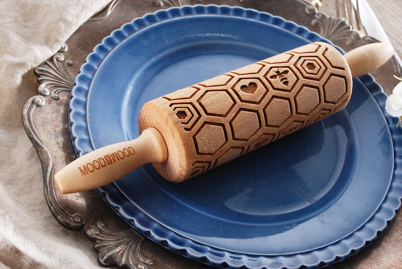 Wooden small rolling pin laser engraved with design of honeycomb, bees, hearts. For embossing cookies. It is lying on a blueplate and metal dish.