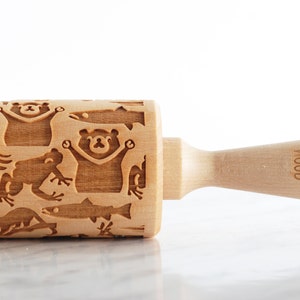 Close-up of a wooden embossed rolling pin engraved with playful animal designs, including bears, fish, and frogs, placed on a smooth, light surface. The handle is visible on the right with subtle branding, while a partially visible cookie with intricate patterns appears on the left edge. Soft lighting enhances the natural wood texture and detailed carvings, creating a clean, minimal, and refined product presentation.