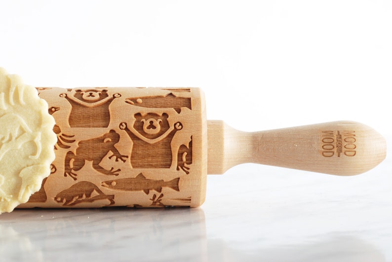 Close-up of a wooden embossed rolling pin engraved with playful animal designs, including bears, fish, and frogs, placed on a smooth, light surface. The handle is visible on the right with subtle branding, while a partially visible cookie with intricate patterns appears on the left edge. Soft lighting enhances the natural wood texture and detailed carvings, creating a clean, minimal, and refined product presentation.