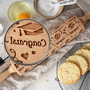 High-quality product photo showing a wooden engraved rolling pin with a detailed graduation-themed pattern, including caps, hearts, and “Congrats” text. A realistic magnifying glass highlights the exact same engraving, revealing deep, precise carving and natural wood grain texture. Surrounding baking elements like eggs, flour, and freshly baked embossed cookies create a warm, artisanal kitchen scene. Soft natural lighting and shallow depth of field emphasize craftsmanship and premium quality.