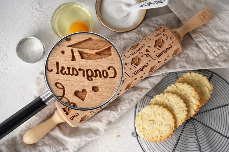 High-quality product photo showing a wooden engraved rolling pin with a detailed graduation-themed pattern, including caps, hearts, and “Congrats” text. A realistic magnifying glass highlights the exact same engraving, revealing deep, precise carving and natural wood grain texture. Surrounding baking elements like eggs, flour, and freshly baked embossed cookies create a warm, artisanal kitchen scene. Soft natural lighting and shallow depth of field emphasize craftsmanship and premium quality.