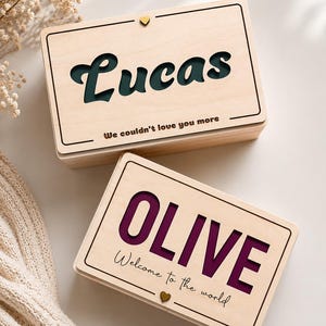 Może przedstawiać: Dwa drewniane pudełka z personalizowanymi imionami. Na górnym pudełku napis "Lucas" w wyciętym wzorze z tekstem "We couldn't love you more." Na dolnym pudełku napis "OLIVE" z tekstem "Welcome to the world."