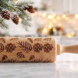 May include: A wooden rolling pin with a decorative pattern of pinecones and leaves. The rolling pin is light brown with a natural wood finish. The handle is engraved with the word "MOOD". The rolling pin is a kitchen tool.