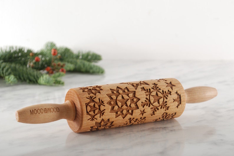 small wooden rolling pin with moodforwood logo embossed with design of different snowflakes. A branch of christmas tree is lying behind it.