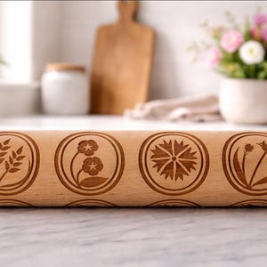 Bright, airy kitchen scene featuring a wooden embossed rolling pin with floral patterns placed on a marble countertop. The background is softly blurred, showing a white ceramic pot filled with fresh flowers, a wooden cutting board, and minimal kitchen elements. Natural light enhances the warm tones of the wood and highlights the intricate engraved details, creating a calm, elegant, and inviting atmosphere.