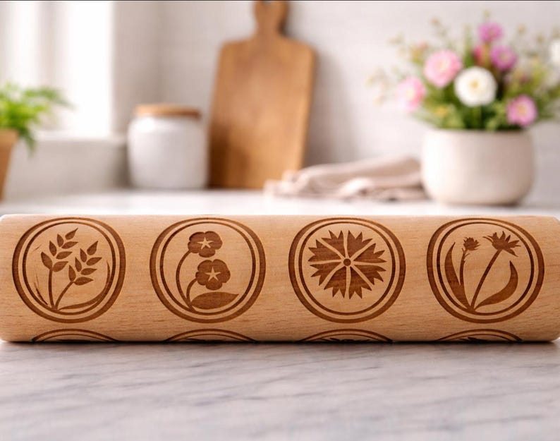 Bright, airy kitchen scene featuring a wooden embossed rolling pin with floral patterns placed on a marble countertop. The background is softly blurred, showing a white ceramic pot filled with fresh flowers, a wooden cutting board, and minimal kitchen elements. Natural light enhances the warm tones of the wood and highlights the intricate engraved details, creating a calm, elegant, and inviting atmosphere.
