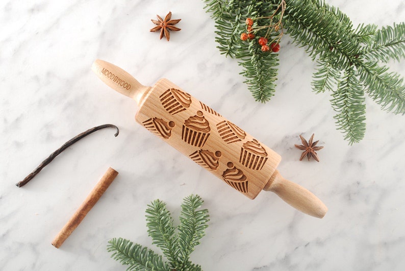 Mini wooden rolling pin with embossing design of cupcakes. It's lying among christmas trees branches and some spices such as vanilla pods, cinnamon stick and  anise seeds.