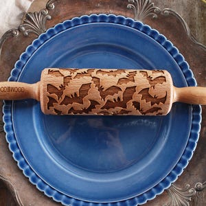 Puede incluir: Un rodillo de madera con un diseño de murciélago grabado en la superficie. El rodillo está sobre un plato azul con un borde plateado. El texto "MOODEWOOD" está grabado en el mango.