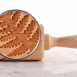 Close-up of a beechwood rolling pin engraved with a detailed fern pattern, placed on a light marble surface. A realistic magnifying glass overlays part of the roller, enlarging the exact same engraving to reveal fine texture, wood grain, and deep carved details. The magnified area clearly shows the precision and depth of the pattern, emphasizing craftsmanship. The handle is visible on the right, and the overall composition is clean, minimal, and styled like high-end product photography.