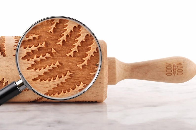 Close-up of a beechwood rolling pin engraved with a detailed fern pattern, placed on a light marble surface. A realistic magnifying glass overlays part of the roller, enlarging the exact same engraving to reveal fine texture, wood grain, and deep carved details. The magnified area clearly shows the precision and depth of the pattern, emphasizing craftsmanship. The handle is visible on the right, and the overall composition is clean, minimal, and styled like high-end product photography.