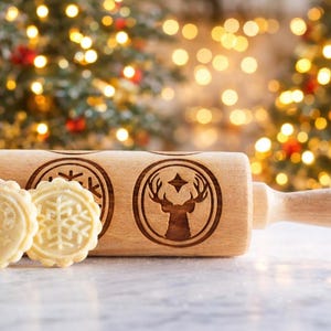 Wooden embossed rolling pin with festive holiday patterns placed on a light marble surface, with three freshly baked cookies in front showing detailed imprints such as holly and a snowflake. In the background, softly blurred Christmas lights and decorated trees create a warm golden bokeh effect. The scene highlights the natural wood texture, precise engraving, and cozy seasonal atmosphere, combining product detail with a festive, high-end lifestyle setting.