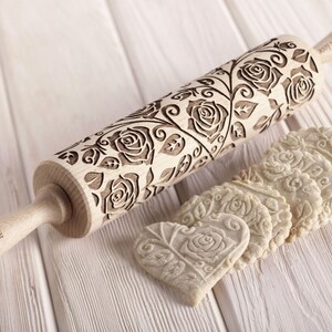 A wooden rolling pin with a floral design featuring roses and leaves. The rolling pin is engraved with the text "Your Wish Will Come True". The rolling pin is shown with several heart-shaped cookies that have been embossed with the same floral design.