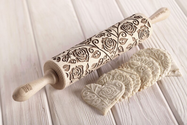 A wooden rolling pin with a floral design featuring roses and leaves. The rolling pin is engraved with the text "Your Wish Will Come True". The rolling pin is shown with several heart-shaped cookies that have been embossed with the same floral design.