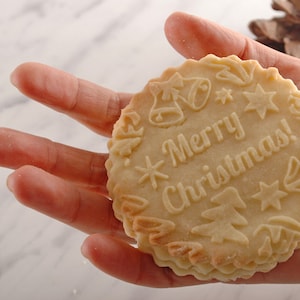 Handholding a round big cookie with merry christmas text embossed on it.