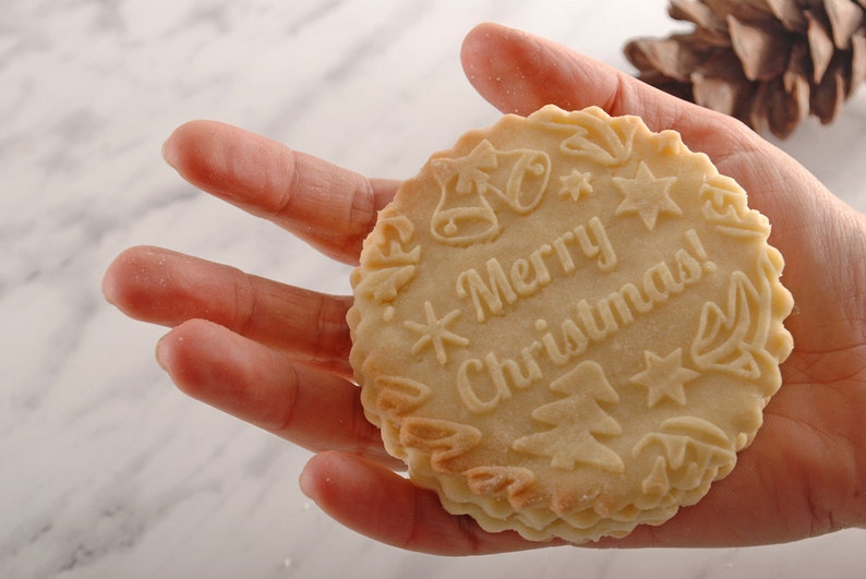 Handholding a round big cookie with merry christmas text embossed on it.