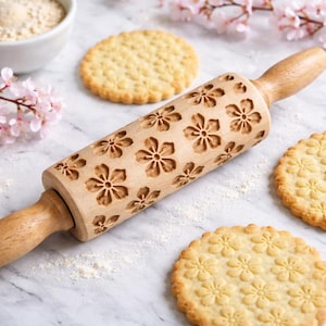 May include: A wooden rolling pin featuring a floral design, with several cookies displaying the same pattern. The rolling pin is light brown, and the cookies are golden. Flour and pink flowers are scattered around the scene, suggesting baking.
