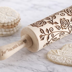 Wooden embossed rolling pin with elegant rose floral pattern resting on a light marble kitchen countertop next to decorative embossed cookies. The engraved beechwood roller creates detailed floral designs in cookie dough, perfect for baking decorative biscuits, fondant or pastry. Romantic baking tool ideal for Valentine cookies, wedding baking, gifts for bakers and homemade cookie decorating.