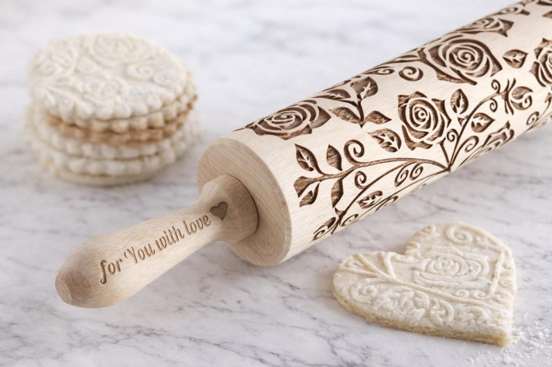 Wooden embossed rolling pin with elegant rose floral pattern resting on a light marble kitchen countertop next to decorative embossed cookies. The engraved beechwood roller creates detailed floral designs in cookie dough, perfect for baking decorative biscuits, fondant or pastry. Romantic baking tool ideal for Valentine cookies, wedding baking, gifts for bakers and homemade cookie decorating.