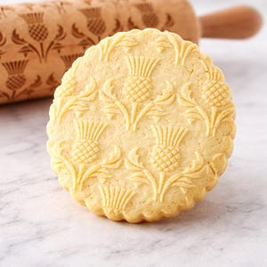Close-up of a large, golden butter cookie with intricate embossed thistle pattern, sharply detailed and centered in the frame. The cookie fills most of the image, highlighting its crisp edges and textured surface. Behind it, a wooden engraved rolling pin with the same pattern is softly blurred, adding context without distraction. The scene is set on a light marble surface with a clean, neutral background. Soft natural lighting enhances the warm tones and depth, creating a premium,
