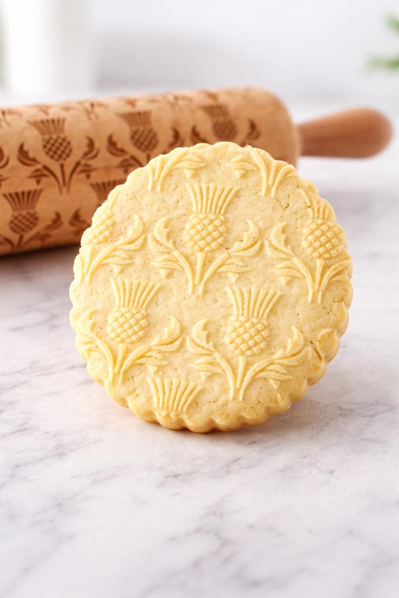 Close-up of a large, golden butter cookie with intricate embossed thistle pattern, sharply detailed and centered in the frame. The cookie fills most of the image, highlighting its crisp edges and textured surface. Behind it, a wooden engraved rolling pin with the same pattern is softly blurred, adding context without distraction. The scene is set on a light marble surface with a clean, neutral background. Soft natural lighting enhances the warm tones and depth, creating a premium,